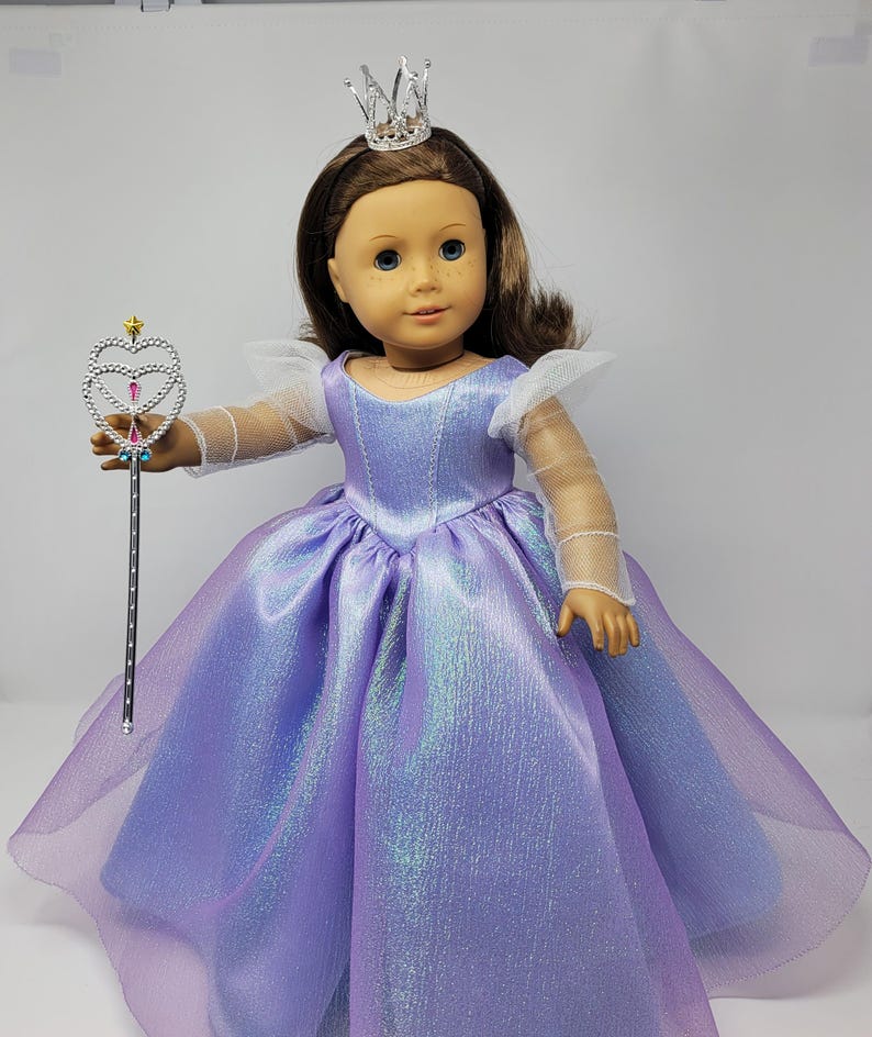 May include: A doll dressed as a princess, wearing a shimmering lavender gown with a sheer overlay. The doll has brown hair, blue eyes, and a silver crown. She holds a silver wand with a heart-shaped top, perfect for imaginative play.
