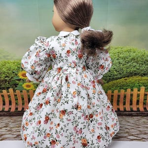 K&ouml;nnte beinhalten: Eine Puppe tr&auml;gt ein wei&szlig;es Kleid mit Blumenmuster, wei&szlig;em Kragen und Puff&auml;rmeln. Das Kleid hat ein Muster aus roten, orangefarbenen und rosa Blumen mit gr&uuml;nen Bl&auml;ttern. Das Kleid hat einen Knopfverschluss auf der R&uuml;ckseite.