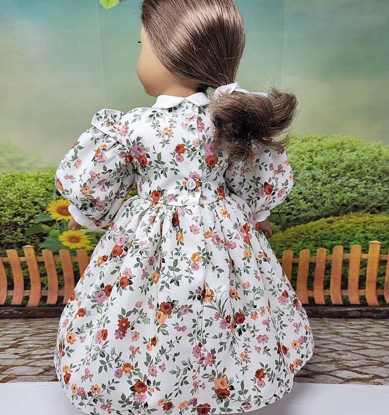 K&ouml;nnte beinhalten: Eine Puppe tr&auml;gt ein wei&szlig;es Kleid mit Blumenmuster, wei&szlig;em Kragen und Puff&auml;rmeln. Das Kleid hat ein Muster aus roten, orangefarbenen und rosa Blumen mit gr&uuml;nen Bl&auml;ttern. Das Kleid hat einen Knopfverschluss auf der R&uuml;ckseite.