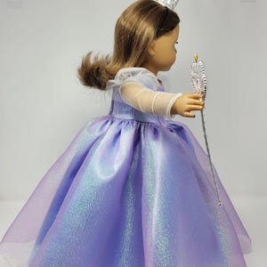 May include: A doll wearing a lavender princess dress with a shimmering, iridescent finish. The dress has a full skirt and sheer sleeves. The doll also wears a silver crown and holds a silver wand with a star-shaped top.