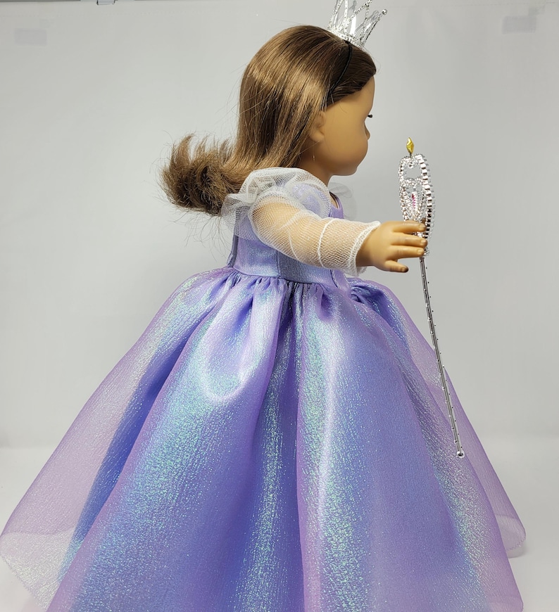 May include: A doll wearing a lavender princess dress with a shimmering, iridescent finish. The dress has a full skirt and sheer sleeves. The doll also wears a silver crown and holds a silver wand with a star-shaped top.