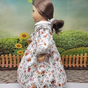 K&ouml;nnte beinhalten: Eine Puppe in einem wei&szlig;en Kleid mit einem Blumenmuster aus orangefarbenen, rosa und roten Blumen. Das Kleid hat einen wei&szlig;en Kragen und lange &Auml;rmel. Die Puppe hat braunes Haar, das zu einem Pferdeschwanz gebunden ist.