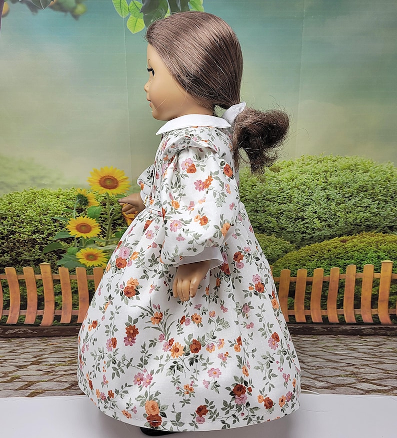 K&ouml;nnte beinhalten: Eine Puppe in einem wei&szlig;en Kleid mit einem Blumenmuster aus orangefarbenen, rosa und roten Blumen. Das Kleid hat einen wei&szlig;en Kragen und lange &Auml;rmel. Die Puppe hat braunes Haar, das zu einem Pferdeschwanz gebunden ist.