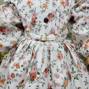 K&ouml;nnte beinhalten: Ein wei&szlig;es Blumenkleid mit einem Muster aus rosa, orangefarbenen und braunen Blumen und gr&uuml;nen Bl&auml;ttern. Das Kleid hat ein eng anliegendes Oberteil mit einem braunen Knopf, eine Taille mit G&uuml;rtel und goldener Schnalle sowie einen plissierten Rock. Die &Auml;rmel sind gepufft.