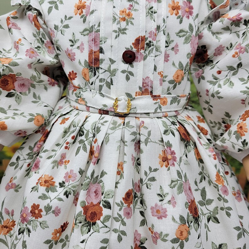 K&ouml;nnte beinhalten: Ein wei&szlig;es Blumenkleid mit einem Muster aus rosa, orangefarbenen und braunen Blumen und gr&uuml;nen Bl&auml;ttern. Das Kleid hat ein eng anliegendes Oberteil mit einem braunen Knopf, eine Taille mit G&uuml;rtel und goldener Schnalle sowie einen plissierten Rock. Die &Auml;rmel sind gepufft.
