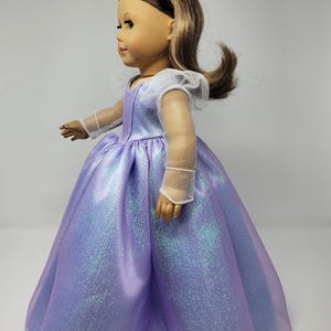 May include: A doll wearing a lavender princess gown with a shimmering, iridescent effect. The dress has a full skirt and sheer sleeves. The doll has brown hair and a silver tiara.
