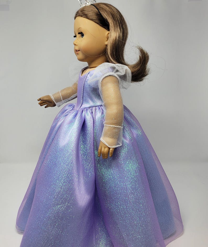 May include: A doll wearing a lavender princess gown with a shimmering, iridescent effect. The dress has a full skirt and sheer sleeves. The doll has brown hair and a silver tiara.
