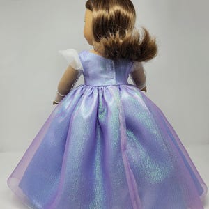 May include: A doll wearing a shimmering lavender princess gown with a full skirt. The dress has a fitted bodice and sheer, iridescent overlay. The doll has brown hair and a silver tiara. The sleeves are white and sheer.