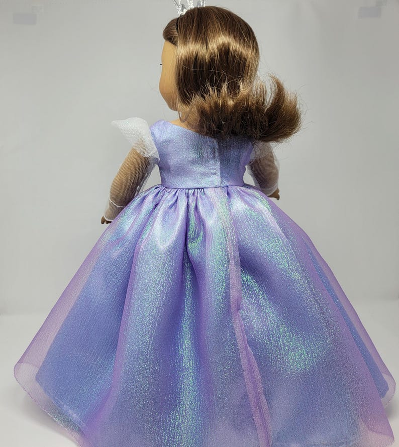 May include: A doll wearing a shimmering lavender princess gown with a full skirt. The dress has a fitted bodice and sheer, iridescent overlay. The doll has brown hair and a silver tiara. The sleeves are white and sheer.
