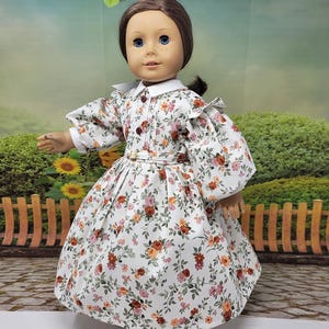K&ouml;nnte beinhalten: Eine Puppe in einem wei&szlig;en Kleid mit Blumenmuster, langen &Auml;rmeln, einem wei&szlig;en Kragen und einem passenden G&uuml;rtel. Das Kleid hat ein Muster aus rosa, orangefarbenen und gr&uuml;nen Blumen. Die Puppe hat braune Haare und blaue Augen.