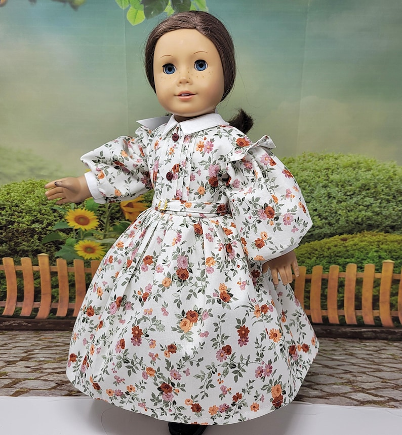 K&ouml;nnte beinhalten: Eine Puppe in einem wei&szlig;en Kleid mit Blumenmuster, langen &Auml;rmeln, einem wei&szlig;en Kragen und einem passenden G&uuml;rtel. Das Kleid hat ein Muster aus rosa, orangefarbenen und gr&uuml;nen Blumen. Die Puppe hat braune Haare und blaue Augen.