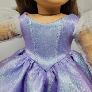 May include: A doll wearing a lavender dress with a shimmering effect. The bodice has a V-neck design with silver stitching. The dress features a full skirt and white, tulle sleeves. The doll's hair is brown.
