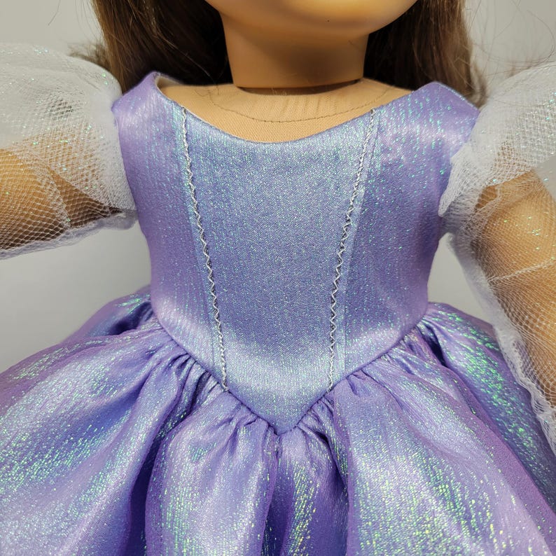 May include: A doll wearing a lavender dress with a shimmering effect. The bodice has a V-neck design with silver stitching. The dress features a full skirt and white, tulle sleeves. The doll's hair is brown.