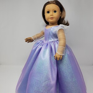 May include: A doll wearing a shimmering lavender princess gown with a full skirt. The doll has brown hair, blue eyes, and a silver crown. The dress features sheer sleeves and a white collar.