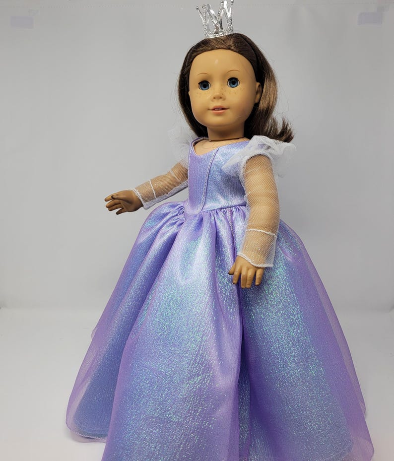 May include: A doll wearing a shimmering lavender princess gown with a full skirt. The doll has brown hair, blue eyes, and a silver crown. The dress features sheer sleeves and a white collar.