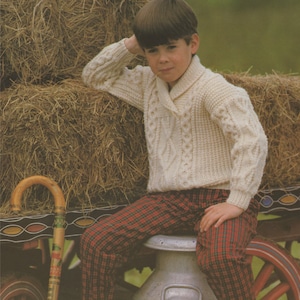 May include: A young person wearing a white cable knit sweater with a shawl collar, red and black plaid pants, and brown shoes. They are sitting on a metal milk can in front of a bale of hay.