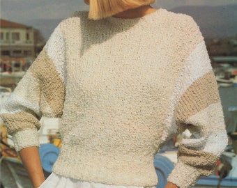 Puede incluir: Un suéter de punto color crema con detalles beige y blancos en las mangas. El suéter se combina con pantalones blancos y un sombrero fedora claro con una banda negra. El estilo general es casual y relajado.