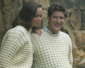 May include: Two people wearing white crocheted sweaters. The woman is wearing blue jeans and the man is wearing blue jeans. They are standing in front of a stone wall.