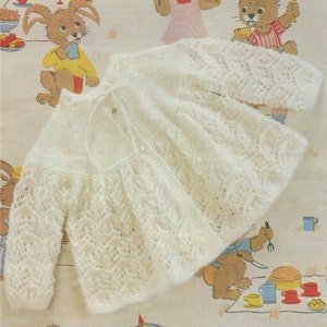 以下が含まれることがあります： レース模様とボタン留めが付いた白いニットのベビーカーディガン。カーディガンは、ウサギ柄の布地に重ねて撮影されています。