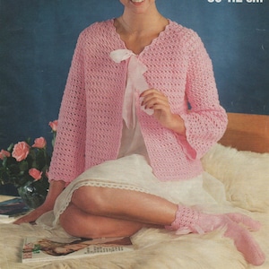 Peut inclure: Un cardigan en crochet rose avec une bordure en dentelle blanche et un nœud rose. Le cardigan est porté sur une robe de dessous blanche. Le mannequin est assis sur un tapis en fourrure blanche. DK 34-44 pouces 86-112 cm