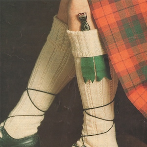 May include: A pair of white knitted socks with black laces, worn with black leather shoes. The socks have green and black embellishments at the top.