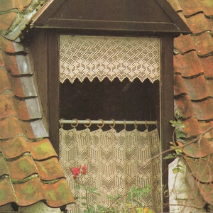 May include: A white crocheted curtain with a scalloped edge hangs in a window. The curtain is made of delicate lace and has a floral pattern. The window is set in a brick wall and is surrounded by a wooden frame.