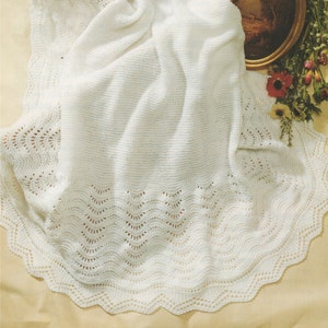 Puede incluir: Una manta de bebé de ganchillo blanco con un delicado borde de encaje. La manta está doblada y drapeada sobre una cesta de mimbre.