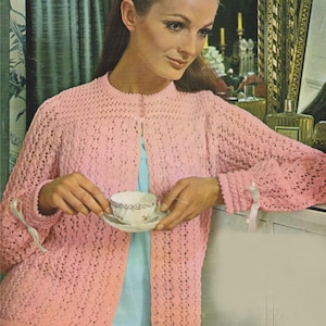May include: A pink crocheted cardigan with a button closure and long sleeves. The cardigan has a delicate openwork pattern and is worn over a light blue top.