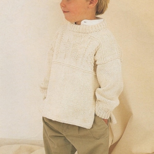 May include: A white knitted jumper with a cable knit pattern. The jumper has a round neckline and long sleeves. The jumper is worn by a child wearing khaki trousers.