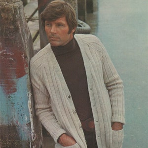 May include: A cream-coloured cardigan with a ribbed texture and a button-up front. The cardigan has two pockets on the front and is worn over a brown turtleneck sweater.