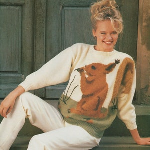 May include: A white knitted jumper with a brown squirrel graphic on the front. The squirrel is holding a nut in its paws and has a bushy tail.