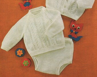 May include: A white knitted baby sweater and matching pants set with a cable knit pattern. The set is laid out on a textured orange surface with two small toy flowers and two toy cats.