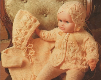 May include: A knitted doll outfit in a pale yellow color. The outfit includes a cardigan, pants, and a bonnet. The outfit is made with a delicate lace pattern.