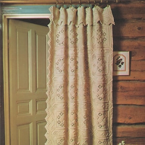 May include: A white crocheted curtain with a diamond pattern hangs in a room with a wooden wall and a door. A vase with a bouquet of pink and white flowers sits in front of the curtain.