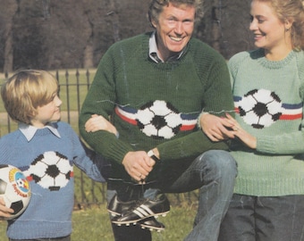 May include: A family of three wearing green, blue, and light green sweaters with a soccer ball design. The sweaters have red, white, and blue stripes across the chest. The family is standing in a park with green grass and trees in the background.