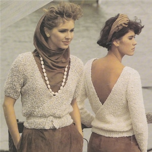May include: Two women wearing white knit sweaters with a V-neck design. The sweater on the left has a brown scarf and a pearl necklace. The sweater on the right has a low back. Both women are wearing brown pants.