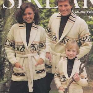 Pode incluir: Três pessoas vestindo cardigãs de tricô cor creme com padrões geométricos marrons e pretos. Os cardigãs têm gola de xale e cinto para amarrar. As pessoas estão em pé em uma área arborizada.