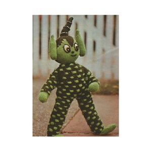 May include: A green knitted toy with brown spots, pointy ears, and a horn. The toy has large eyes and a smiling face.