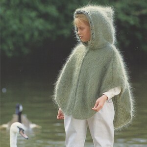 Pode incluir: Uma pessoa jovem vestindo um poncho de malha verde claro com capuz e textura felpuda. O poncho tem uma borda branca ao redor do capuz. A pessoa está vestindo calças brancas e botas cor-de-rosa.