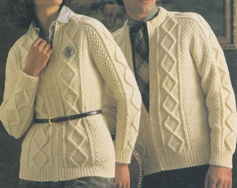 May include: Cream-colored knit zip-up sweaters with diamond and cable patterns. The woman wears a belt and a brooch. The man wears a patterned tie. The sweaters have a classic, textured design.