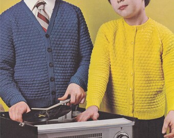May include: Two children, one wearing a blue cardigan and the other wearing a yellow cardigan, are standing next to a record player. The child in the blue cardigan is holding a record and a stylus. The record player has a black base and a silver top with a speaker grill.