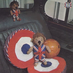 May include: Two knitted lion toys wearing red, white, and blue uniforms with the text "World Cup" on the chest. The toys are sitting on red, white, and blue striped cushions. A soccer ball is next to the larger lion toy.