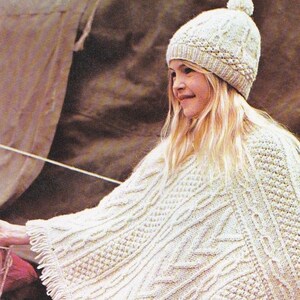 Peut inclure: Un poncho blanc en tricot avec une bordure en franges. Le poncho est à motif de tricot torsadé et est porté par une jeune fille aux cheveux blonds portant un bonnet blanc en tricot avec un pompon.