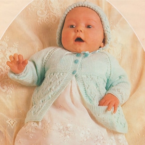 以下が含まれることがあります： 薄いブルーのニットカーディガン、白いドレス、それに合わせたブーティを履いた赤ちゃん。赤ちゃんはレース模様の薄い茶色の表面に寝転んでいます。