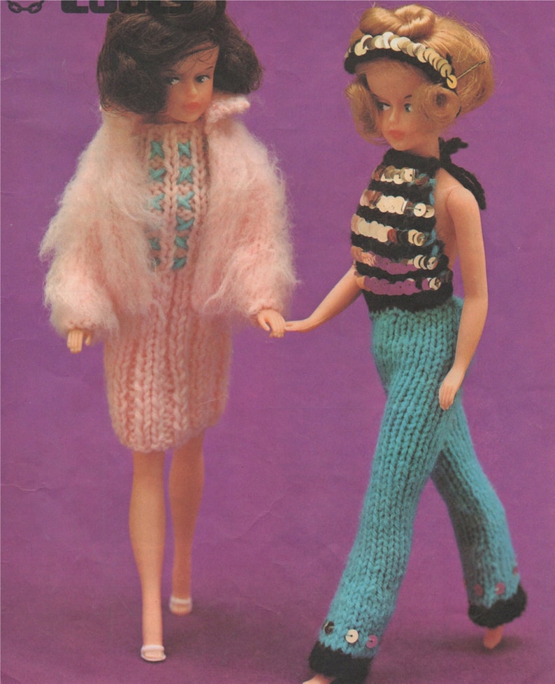 K&ouml;nnte beinhalten: Zwei Puppen in gestrickten Outfits. Die Puppe links tr&auml;gt ein rosa Kleid und eine Jacke. Die Puppe rechts tr&auml;gt ein blaues und schwarzes Halstuch und eine blaue Hose.