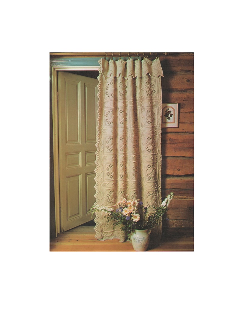 May include: A crocheted curtain with a geometric pattern hangs in a doorway. The curtain is cream colored and has a scalloped edge. A vase of flowers sits on the floor in front of the curtain.