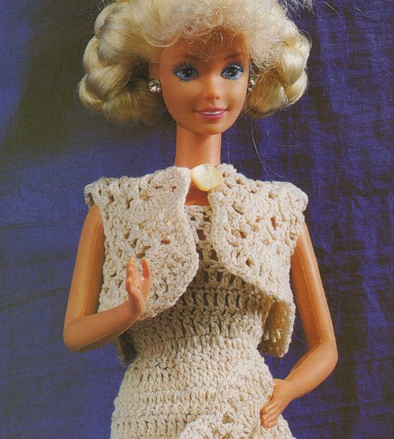May include: A crocheted dress and matching bolero for a doll. The dress and bolero are cream colored and have a delicate floral pattern. The doll has blonde hair and blue eyes.