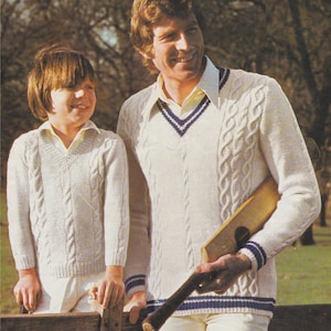 May include: Two people wearing white cable knit sweaters with blue and white trim. The person on the right is holding a wooden cricket bat.