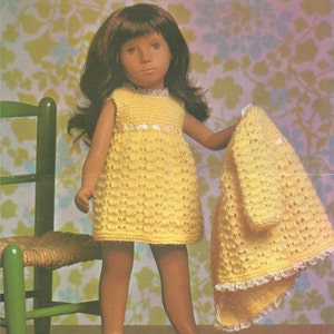 May include: A doll wearing a yellow knitted dress and matching cardigan with white lace trim. The doll has brown hair and brown eyes. The doll is standing in front of a chair with a woven seat.