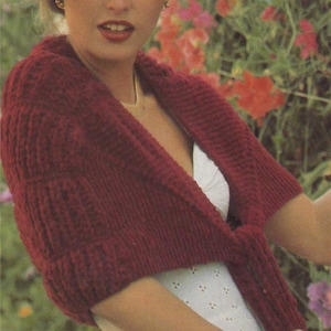 May include: A woman wearing a red knitted shawl over a white eyelet dress. The shawl is tied at the front and has a V-neckline. The woman is standing in front of a background of pink and white flowers.